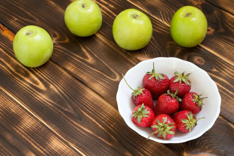 green apples, red strawberries in white bowl on dark wooden table background with fresh fruits and berries with copy space place for text raw foods natural organic food concept summer harvest