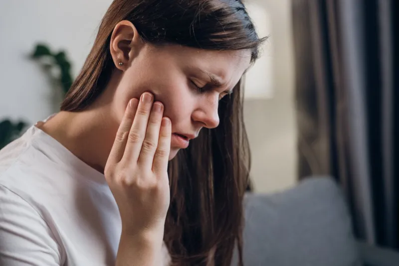 close up of upset young female 20s touching mouth with hand, painful expression because dental illness, dentist concept, sick unhappy brunette woman with caries and toothache sitting on sofa at home
