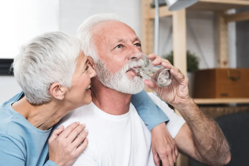 happy beautiful senior man, white beard and grey hair, holding transparent glass in his hand and drink water smiling wife near by concept healthy life and diet for senior people closeup