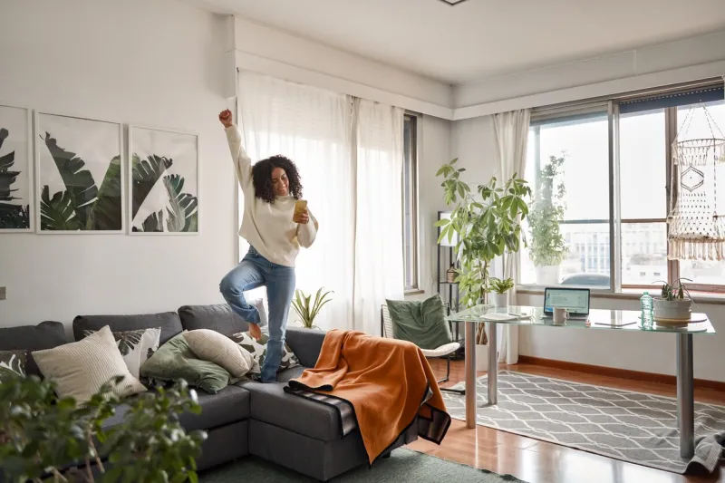 happy excited young woman using smartphone dancing at home latin girl jumping on couch listening music on mobile phone, winning money in app game celebrating standing in living room interior