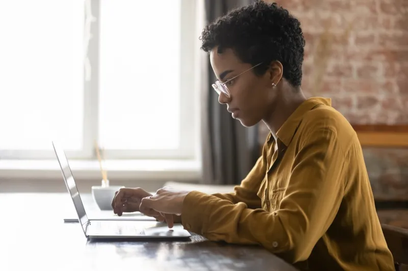 side view focused young african american businesswoman in eyewear working on computer, sitting at table in modern loft office room, typing message, preparing electronic document or presentation