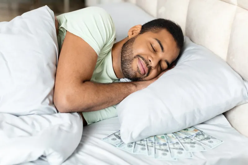 happy peaceful rich africna american man sleeping with bunch of cash dollar banknotes under his pillow in bed, keeping his money savings by him at home, closeup finance management concept