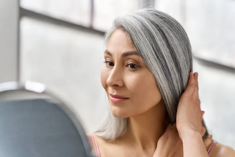 portrait of beautyful happy middle aged mature asian woman, senior older 50s lady pampering touching gray hair looking at herself at mirror indoors ads of lifting anti wrinkle skin hair care spa