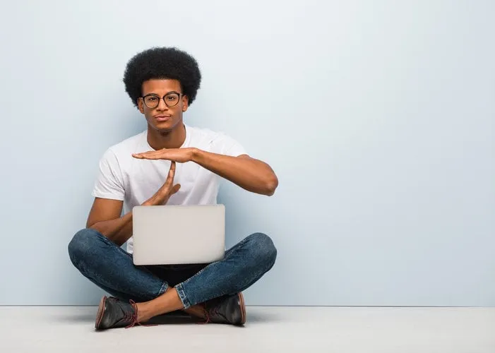 young black man sitting on the floor with a laptop doing a timeout gesture