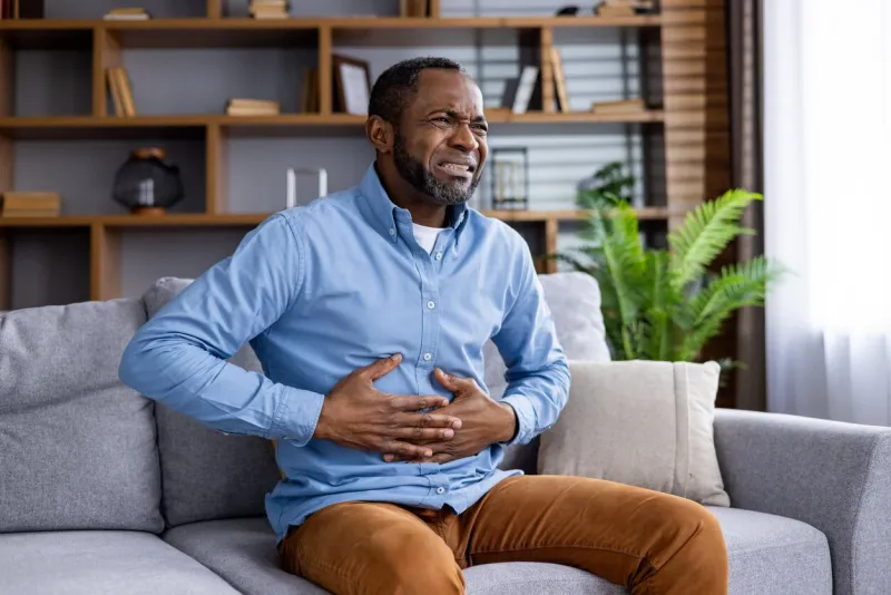 man experiencing abdominal pain while seated on sofa in living room man clutching stomach due to feeling of discomfort concept of health, pain, and discomfort