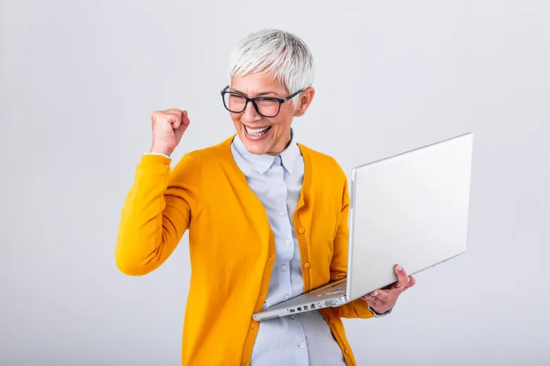 portrait of a cheerful mature woman with a laptop computer and celebrating success isolated over gray background senior lady watching celebrating online bid bet win or great result victory concept