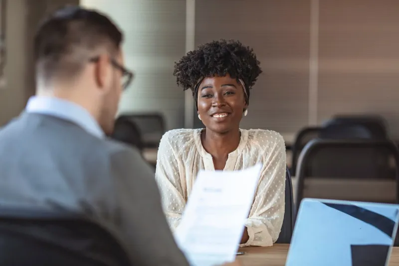 portrait of joyful woman smiling and holding resume while sitting in front of businessman during corporate meeting or job interview - business, career and placement concept