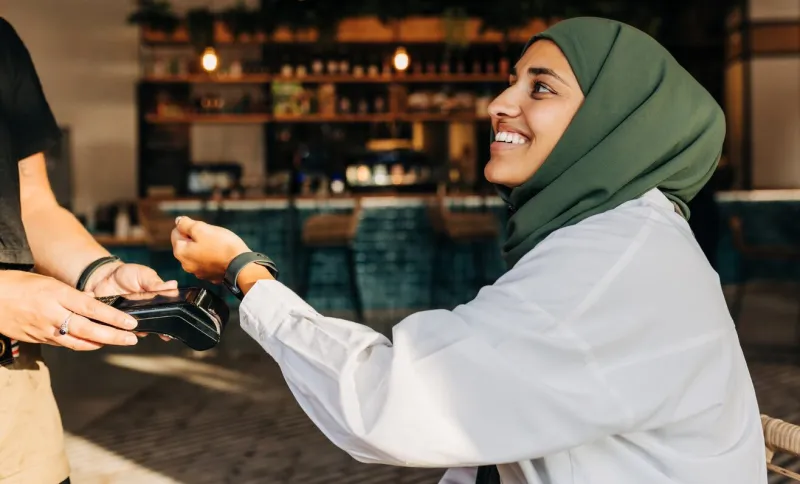 happy muslim woman scanning her smartwatch on a card machine to pay her bill in a cafe woman with a hijab smiling cheerfully while doing a contactless transaction using nfc technology
