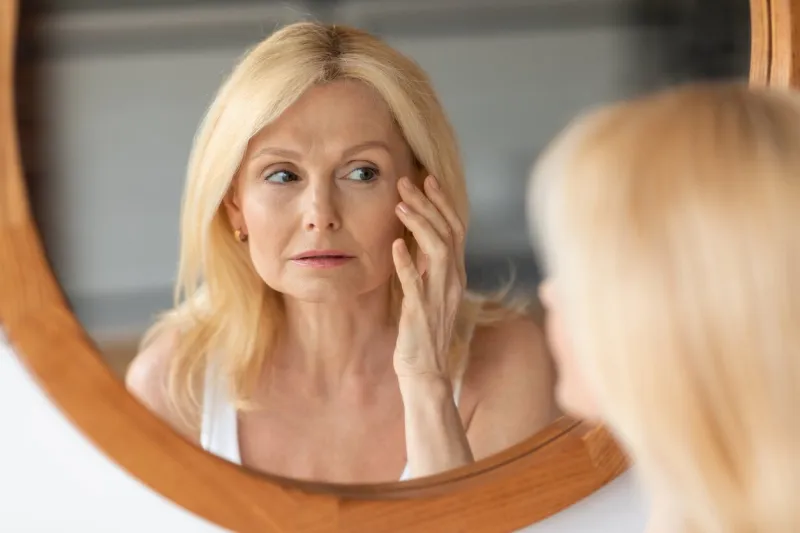 skin aging concept beautiful aged woman looking at mirror at her wrinkles near eyes, checking face, noticing age changes during daily beauty routine at home