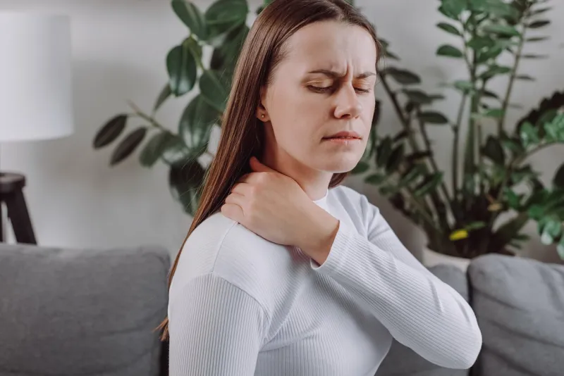 close up of tired unhappy young woman feeling exhausted and suffering from neck pain, sad unhealthy caucasian doing massage sitting alone on grey couch at home health concept