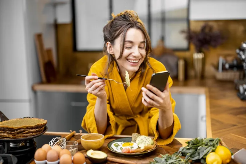 young happy woman dressed in yellow bathrobe enjoys healthy breakfast and reading on phone at home morning affairs and routine