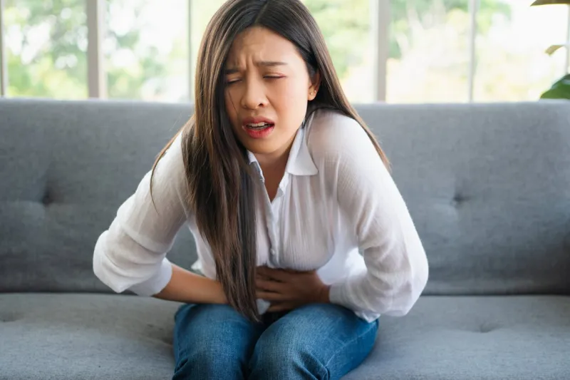 unhappy asian woman sitting on the sofa and holding on stomach suffering abdominal pain that comes from menstruation, diarrhea, or indigestion sickness and healthcare concept