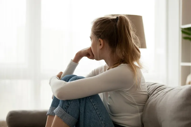 side view young woman looking away at window sitting on couch at home frustrated confused female feels unhappy problem in personal life quarrel break up with boyfriend or unexpected pregnancy concept