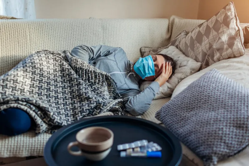 sick woman having flu or cold girl lying in bed having headache wearing protective mask by pills and water on table