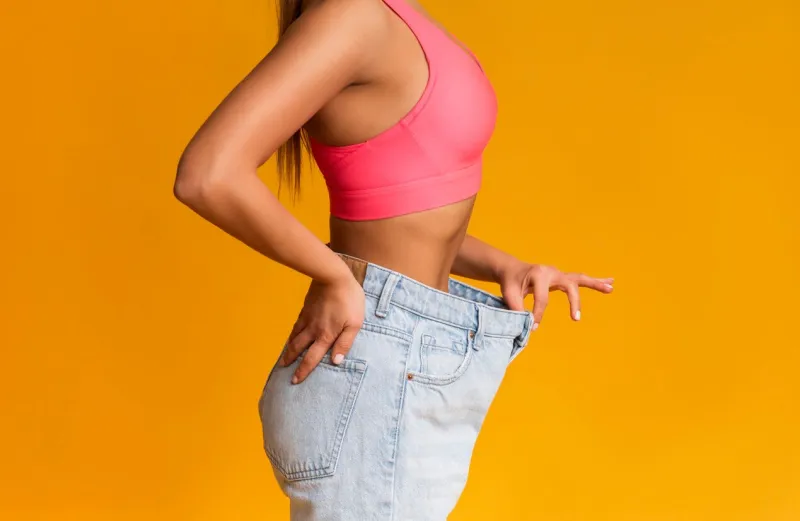 weight loss concept profile portrait of fit girl posing in oversize jeans over yellow background, cropped image with copy space