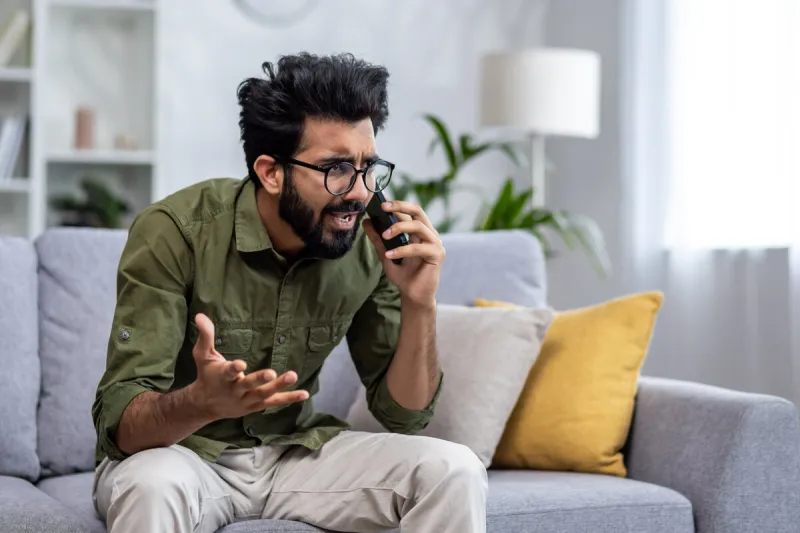 angry and nervous man talking on the phone sitting on the sofa in the living room, hispanic man yelling at the interlocutor on the smartphone, dissatisfied with the service, the customer complains