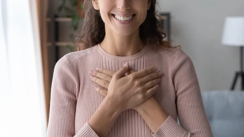 close up focus on happy sincere female holding folded hands on chest emotional positive kind candid millennial woman feeling thankful indoors, showing gratitude sign, believe faith charity concept
