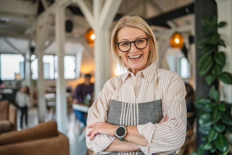 portrait of a happy mature woman in apron stand at restaurant arm crossed, beautiful woman owner stand and smile at restaurant