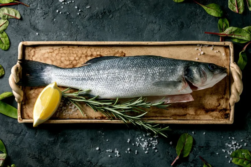 raw fresh uncooked fish sea bass on ceramit plate ready to cook with lemon, sea salt, rosemary, green salad over dark texture concrete background flat lay, space