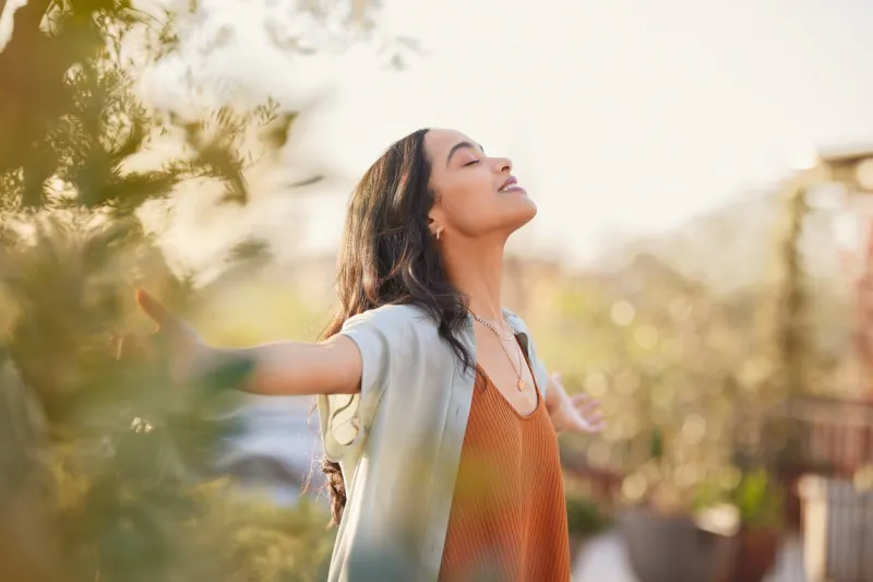 young latin woman with arms outstretched breathing in fresh air during sunrise at the balcony healthy girl enjoying nature while meditating during morning with open arms and closed eyes mindful woman enjoying morning ritual while relaxing in outdoor park