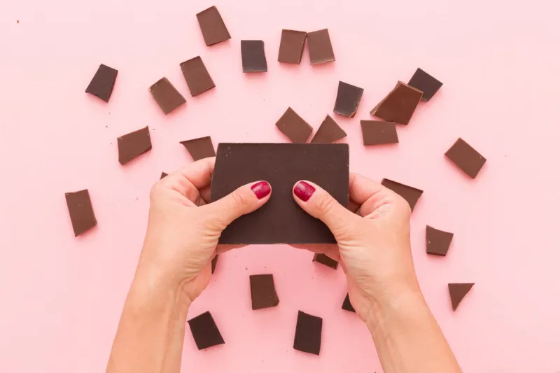 top view on woman's hands breaking a piece of dark chocolate above pink table background