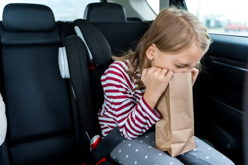 little girl is driving in car kid is sick, feels bad and vomiting into paper bag traveling, riding on road in safe baby seats with child belts fun family trip, activity with parents