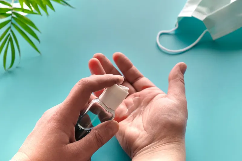 hands of senior female with sanitizer, hand soap mint green background with face mask, pills, natural palm tree leaf social isolation, hygiene, prophylactic measures to fight novel coronavirus