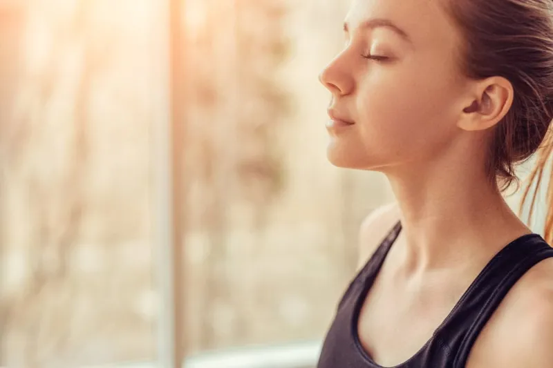 side view of young female with closed eyes breathing deeply while doing respiration exercise during yoga session in gym