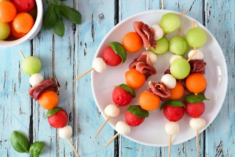 plate of delicious summer fruit skewers with melon, cheese and prosciutto on a rustic blue wood background