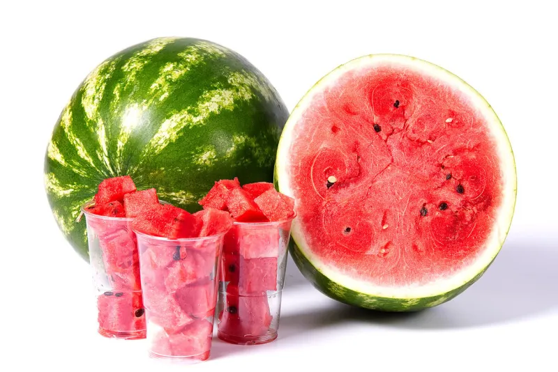 sliced red watermelon cubes in plastic cup a front of fresh organic green watermelon and sliced half of watermelon with red texture