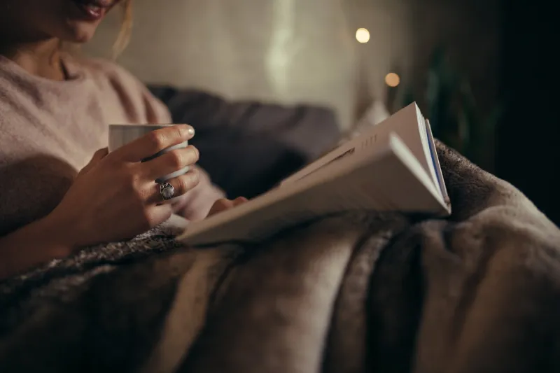 cropped shot of female hands with book and coffee female reading book on bed at night