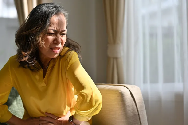 senior woman holding belly, feeling discomfort, unhappy older female sitting on couch, suffering from stomachache, food poisoning, gastritis, abdominal pain