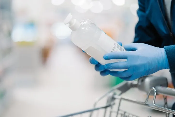 unrecognizable person in medical gloves holds bottle of sanitizer gel, poses in shop, leans at shopping cart, buys product for coronavirus protection, blurred background disinfection, prevention