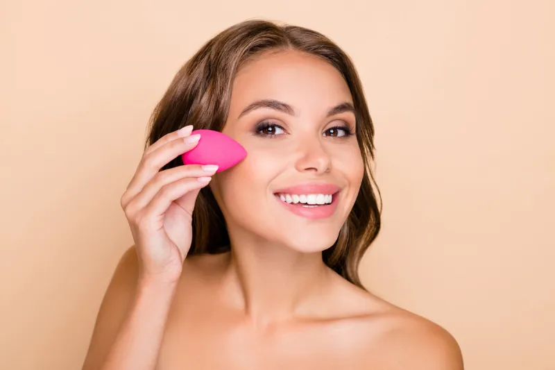 photo of young beautiful smiling woman applying foundation on cheek with beauty blender isolated on beige color background