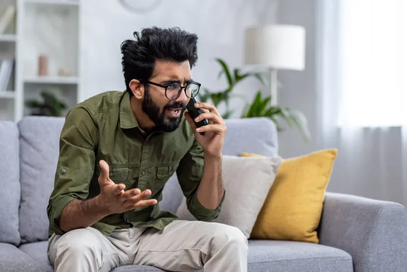 angry and nervous man talking on the phone sitting on the sofa in the living room, hispanic man yelling at the interlocutor on the smartphone, dissatisfied with the service, the customer complains