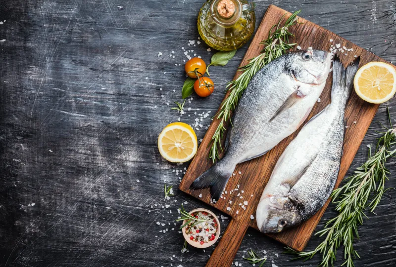 fresh uncooked dorado or sea bream fish with lemon, herbs, olive oil and spices over black backdrop, top view with copy space