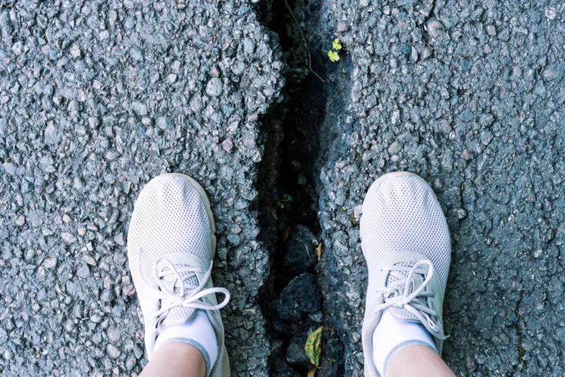 view of two feet in white sneakers standing at the edge of a deep crack in pavement the scene highlights the condition of worn infrastructure and potential hazards