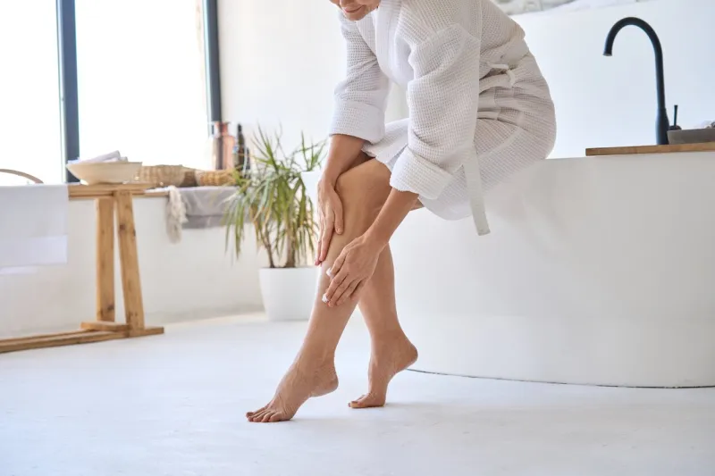 mid age adult 50s age mature woman applying varicose prevention treatment cream massaging legs sitting on bathtub wearing white bathrobe feminine health care after menopause concept