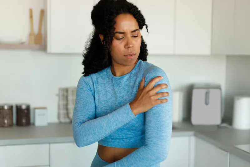 sporty black woman putting her hand on injury shoulder, having pain, standing in kitchen interior at home, resting after domestic training sport concept