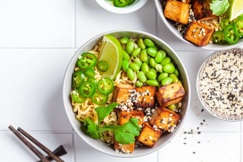 vegan noodles ramen soup with marinated tofu, edamame beans and hot peppers in a gray bowl on a white tile background, top view
