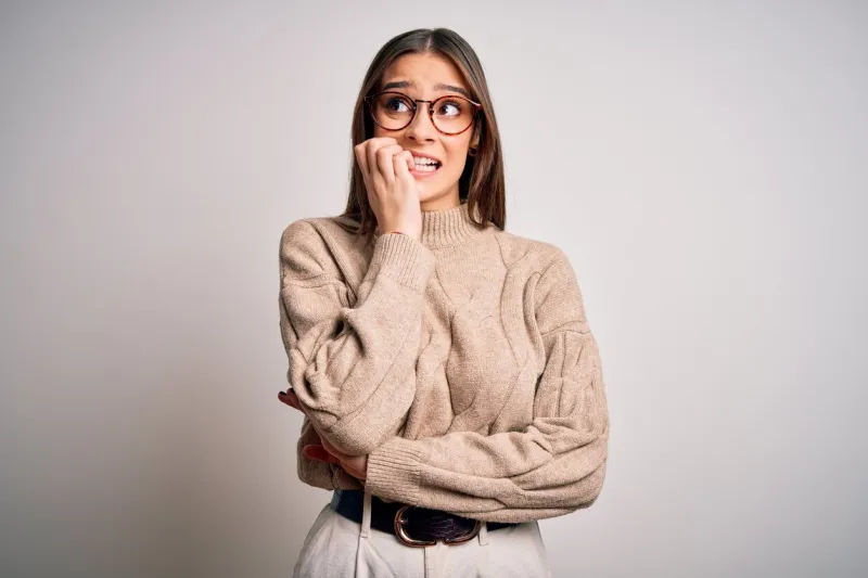 young beautiful brunette businesswoman wearing casual sweater and glasses standing looking stressed and nervous with hands on mouth biting nails anxiety problem