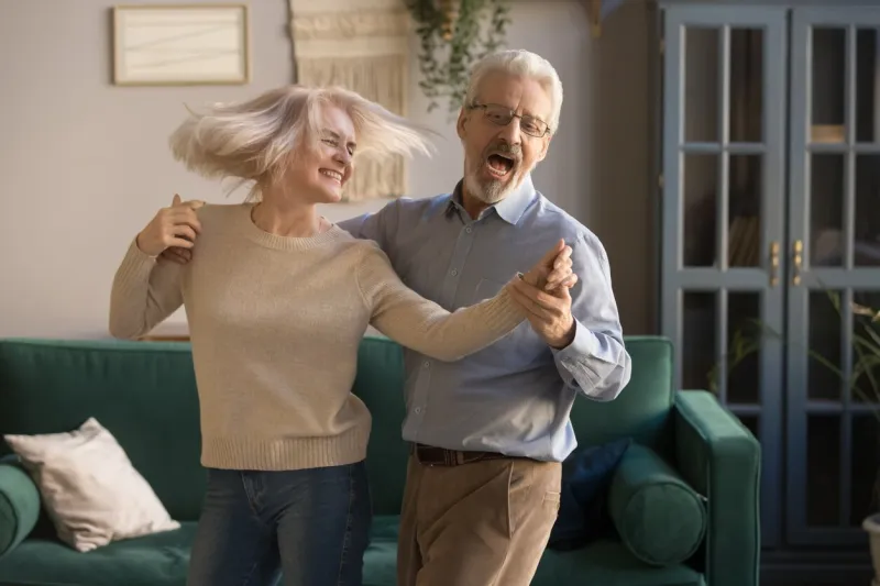 carefree happy active old senior couple dancing jumping laughing in living room, cheerful retired elder husband holding hand of mature middle aged wife enjoy fun leisure retirement lifestyle at home