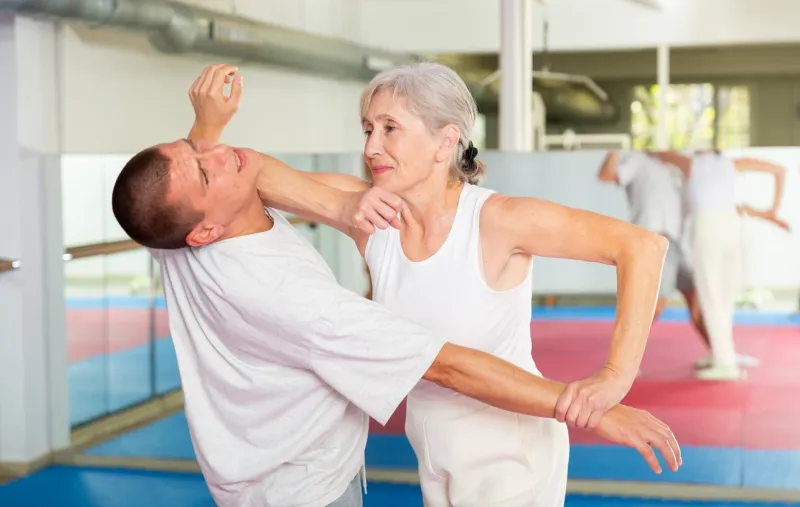 determined aged woman learning self defence techniques in sparring with young man, practicing elbow blow with wristlock to opponent in gym