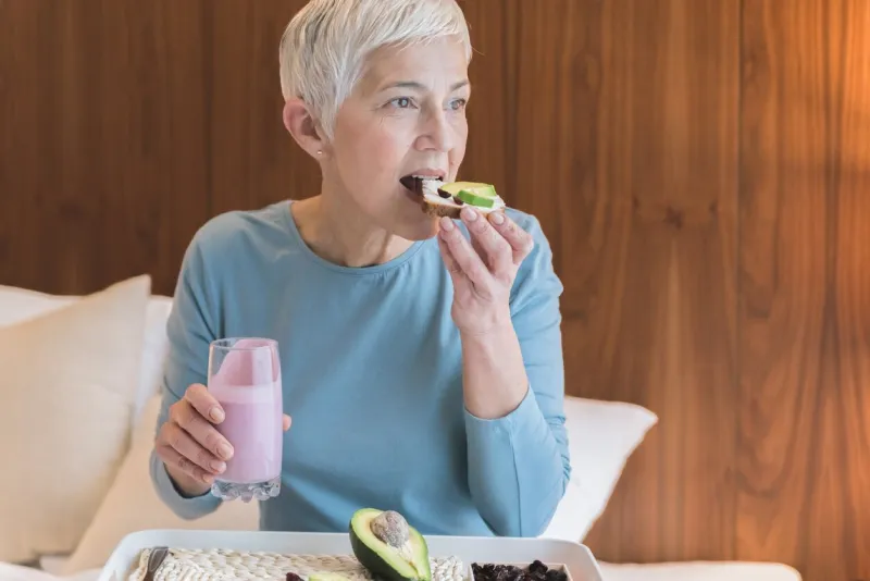 beautiful senior woman eating nutritious healthy toast with avocado and holding fruit yogurt, healthy nutrition concept