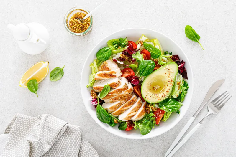 grilled chicken meat and fresh vegetable salad of tomato, avocado, lettuce and spinach healthy and detox food concept ketogenic diet buddha bowl dish on white background, top view
