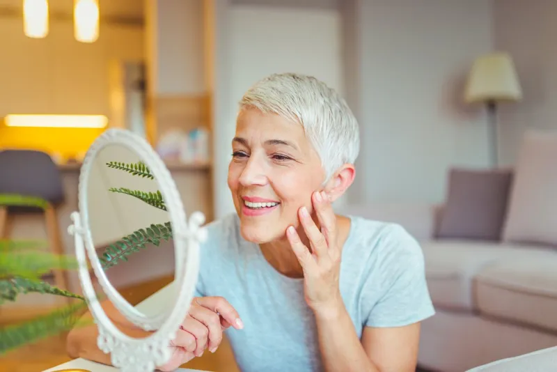 happy mature woman admiring herself in the mirror middle aged woman looking at wrinkles in mirror plastic surgery and collagen injections makeup good skincare habits will have you looking younger