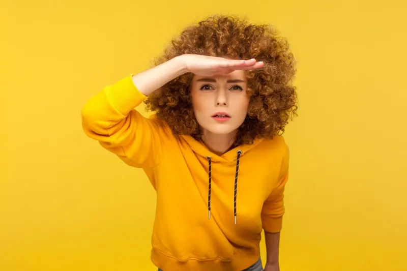 portrait of curious curly-haired woman in urban style hoodie holding hand above eyes and peering into distance, looking far away, expecting and searching someone on horizon studio shot isolated