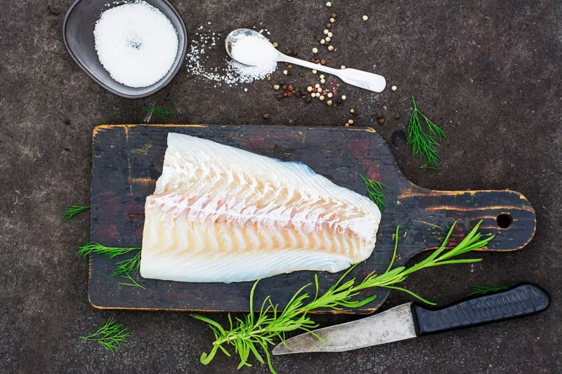 raw cod before cooking on a black chopping board with herbs and sea salt on a dark background top view