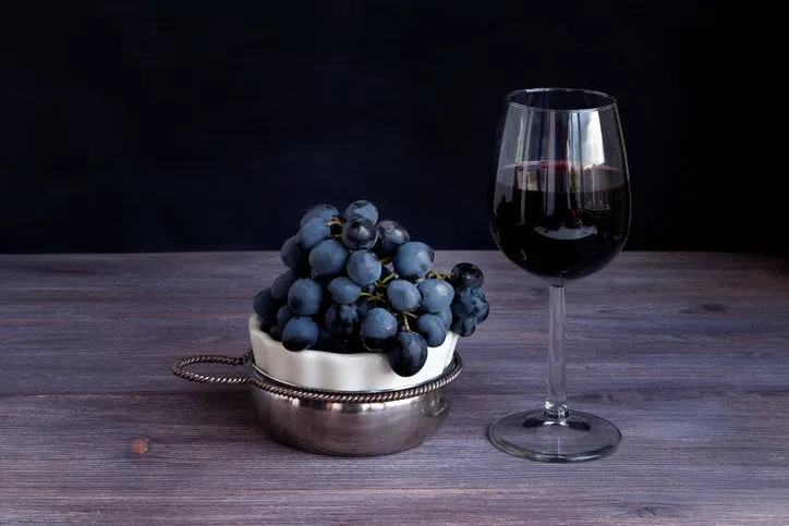 red wine in wine glass and bunch of dark red purple grapes on wooden board against black background toned image copy space