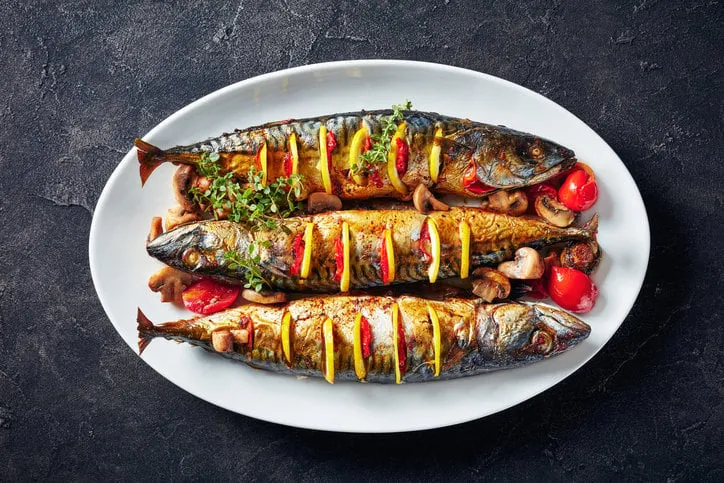 close-up of three whole broiled mackerels with lemon, tomatoes, mushrooms, spices and herbs on a white oval dish, horizontal view from above, flatlay